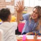 A preschool child sits at a table with his teacher as the two high-five.