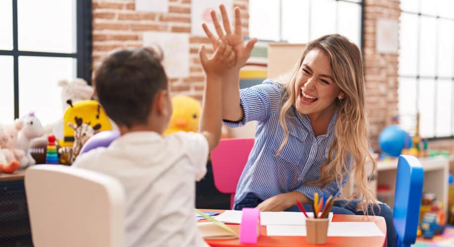 A preschool child sits at a table with his teacher as the two high-five.