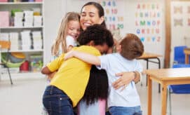 A group of preschool children hug their teacher in an early childhood education classroom.