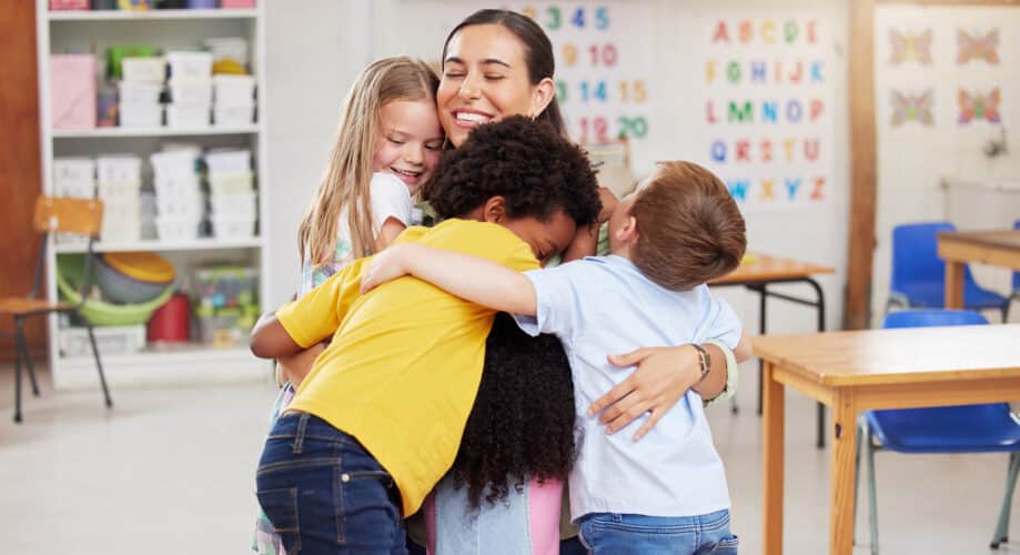 A group of preschool children hug their teacher in an early childhood education classroom.