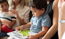 Two toddler boys sit on their caregivers’ laps while one explores a children’s book about crocodiles.