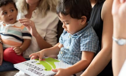 Two toddler boys sit on their caregivers’ laps while one explores a children’s book about crocodiles.