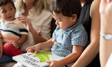 Two toddler boys sit on their caregivers’ laps while one explores a children’s book about crocodiles.