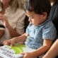 Two toddler boys sit on their caregivers’ laps while one explores a children’s book about crocodiles.