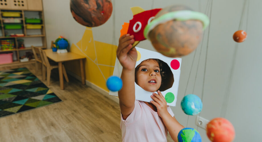 Preschool girl plays with a hanging solar system display in her early learning classroom.