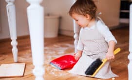 A toddler child sits on the floor with a small broom and dustpan, helping to clean up in an early learning classroom.