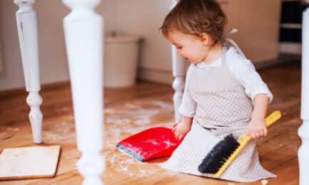 A toddler child sits on the floor with a small broom and dustpan, helping to clean up in an early learning classroom.