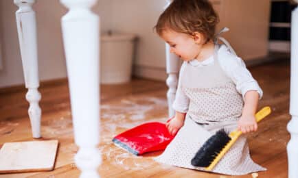 A toddler child sits on the floor with a small broom and dustpan, helping to clean up in an early learning classroom.