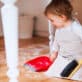 A toddler child sits on the floor with a small broom and dustpan, helping to clean up in an early learning classroom.