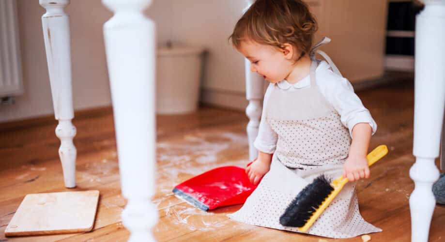 A toddler child sits on the floor with a small broom and dustpan, helping to clean up in an early learning classroom.