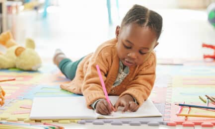 A young girl lies on the floor of a preschool classroom while she happily draws in her journal.