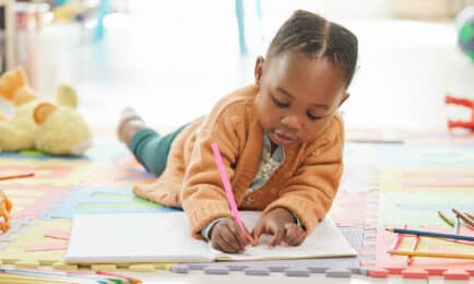 A young girl lies on the floor of a preschool classroom while she happily draws in her journal.