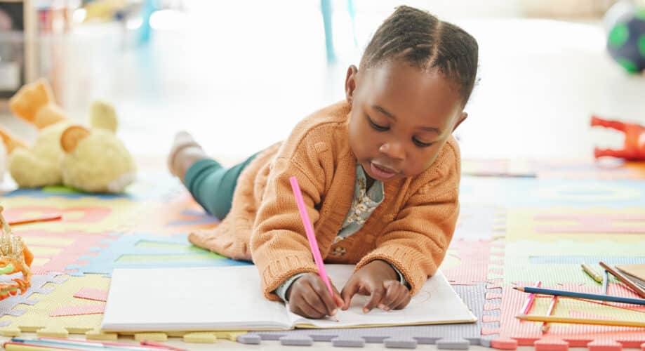 A young girl lies on the floor of a preschool classroom while she happily draws in her journal.