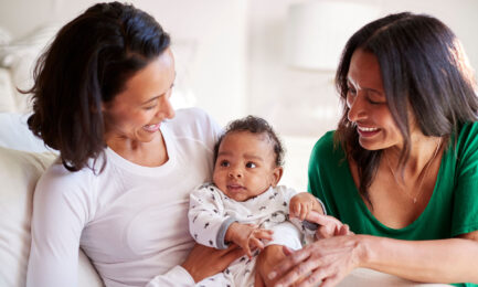 A baby boy sits on his mother’s lap, alongside another caregiver.