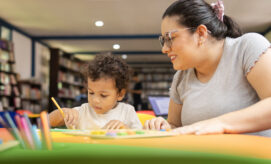 A young boy sits at a table and colors with markers while a caregiver provides support.