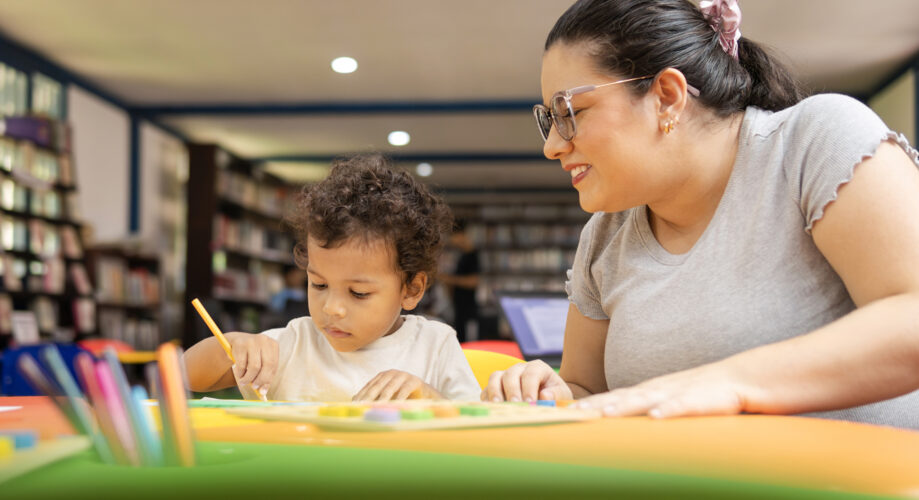A young boy sits at a table and colors with markers while a caregiver provides support.