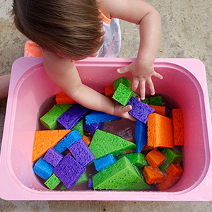 Toddler searching through a bin of sponges cut into basic geometric shapes.