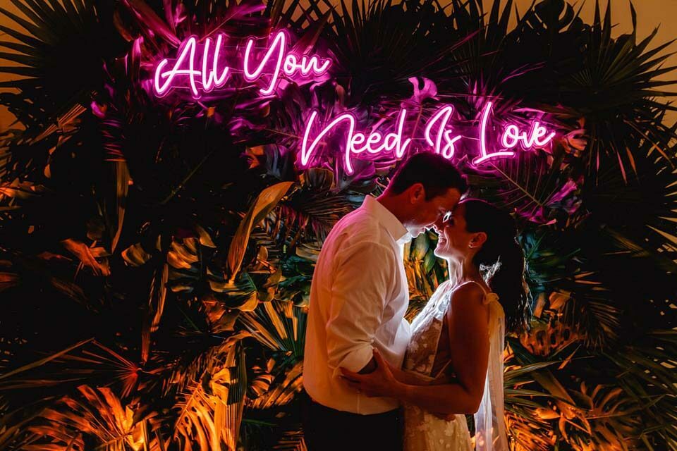 Bride and groom pose head-to-head in front of a vivid pink All You Need Is Love neon sign
