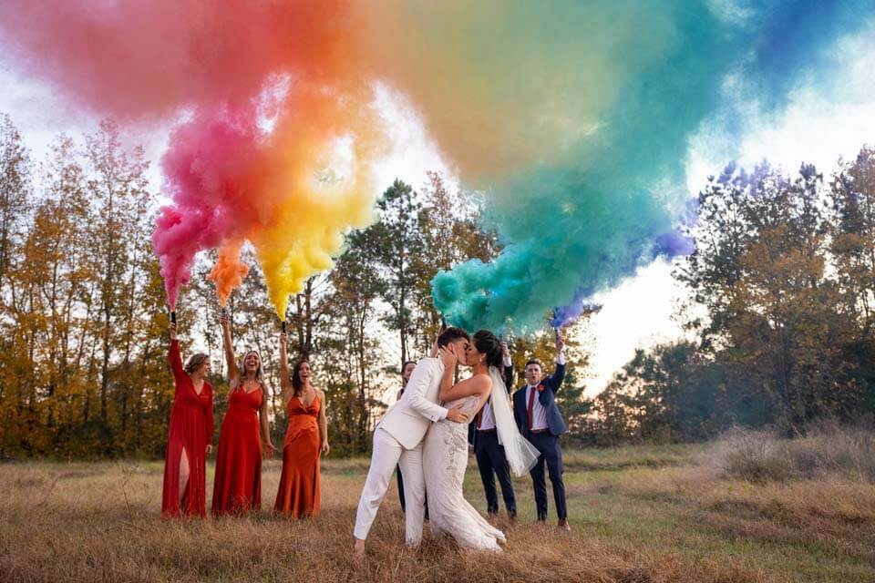 Two brides kiss as their wedding party shoots off a rainbow cloud of smoke bombs