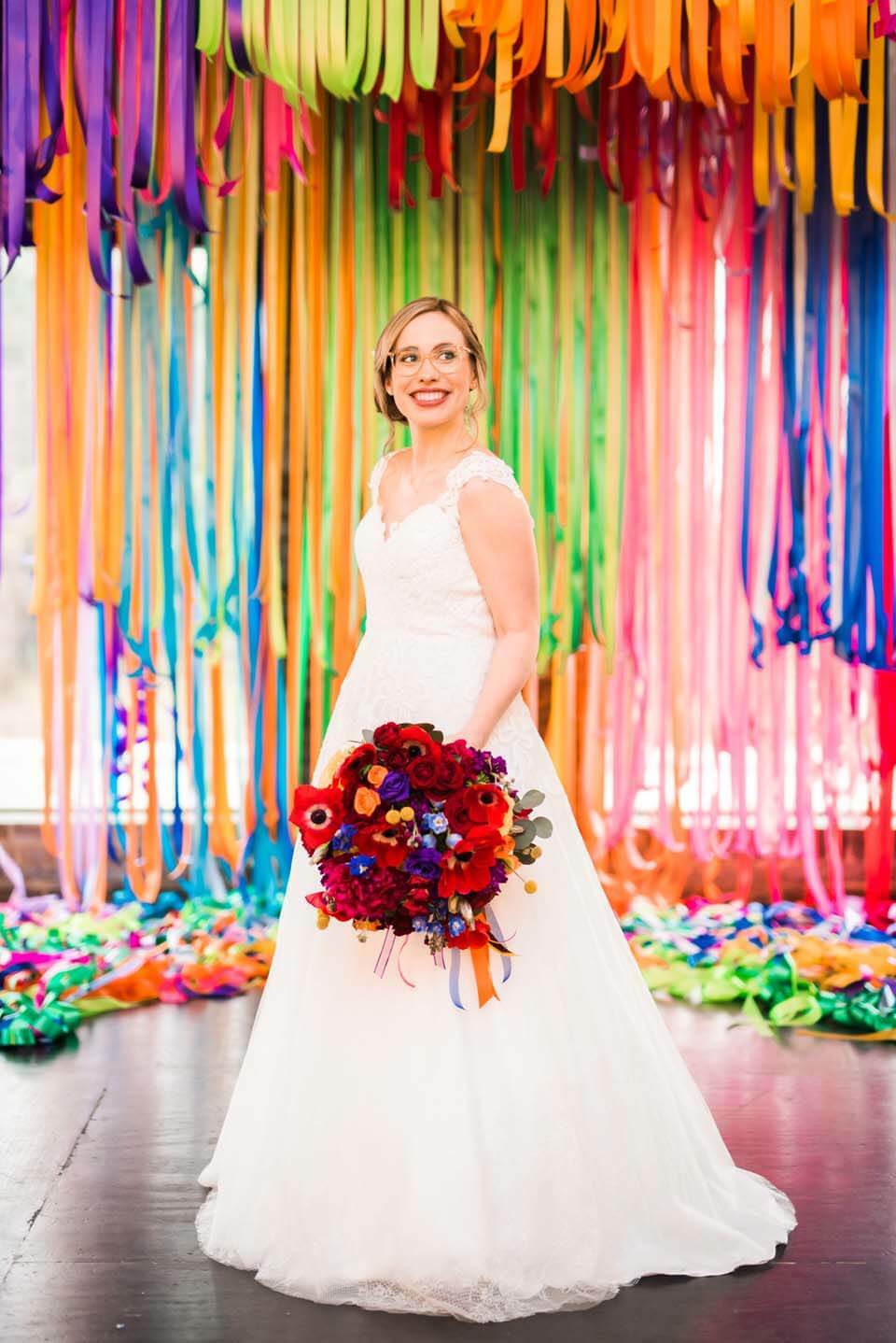 A bride wearing glasses and holding a lush bouquet poses in front of a backdrop of hanging rainbow streamers