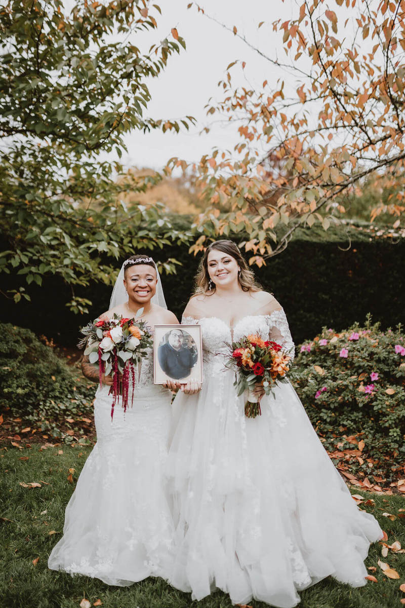 Two brides hold a framed photo of a loved one