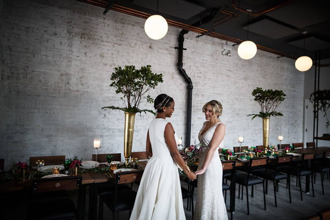Two brides holding hands in front of their industrial-chic wedding reception