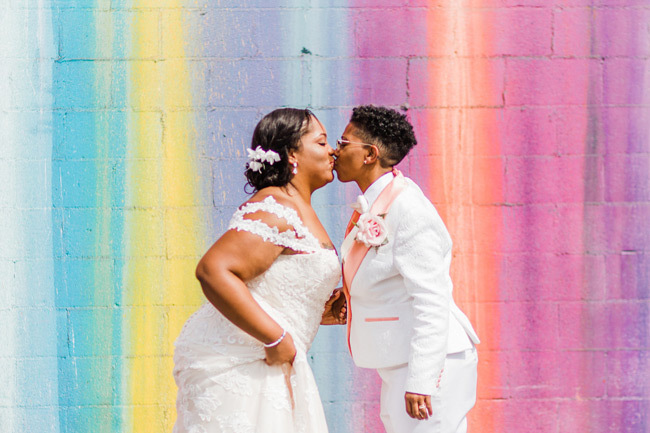 Lesbian Wedding Ideas | Two brides kiss in front of a rainbow wall Lesbian Wedding Ideas | Two brides kiss in front of a rainbow wall