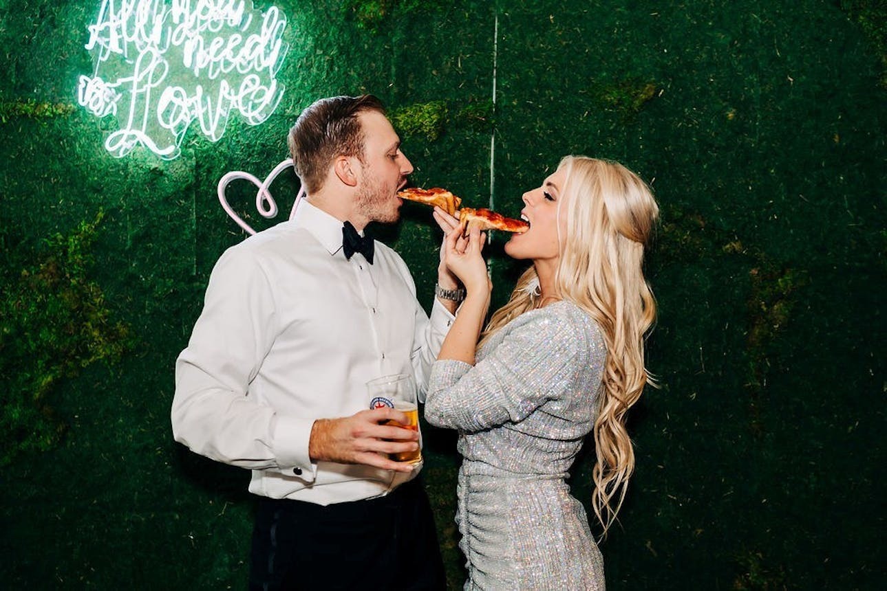 Newlyweds eating pizza in front of a neon sign | Memorable rehearsal dinner ideas