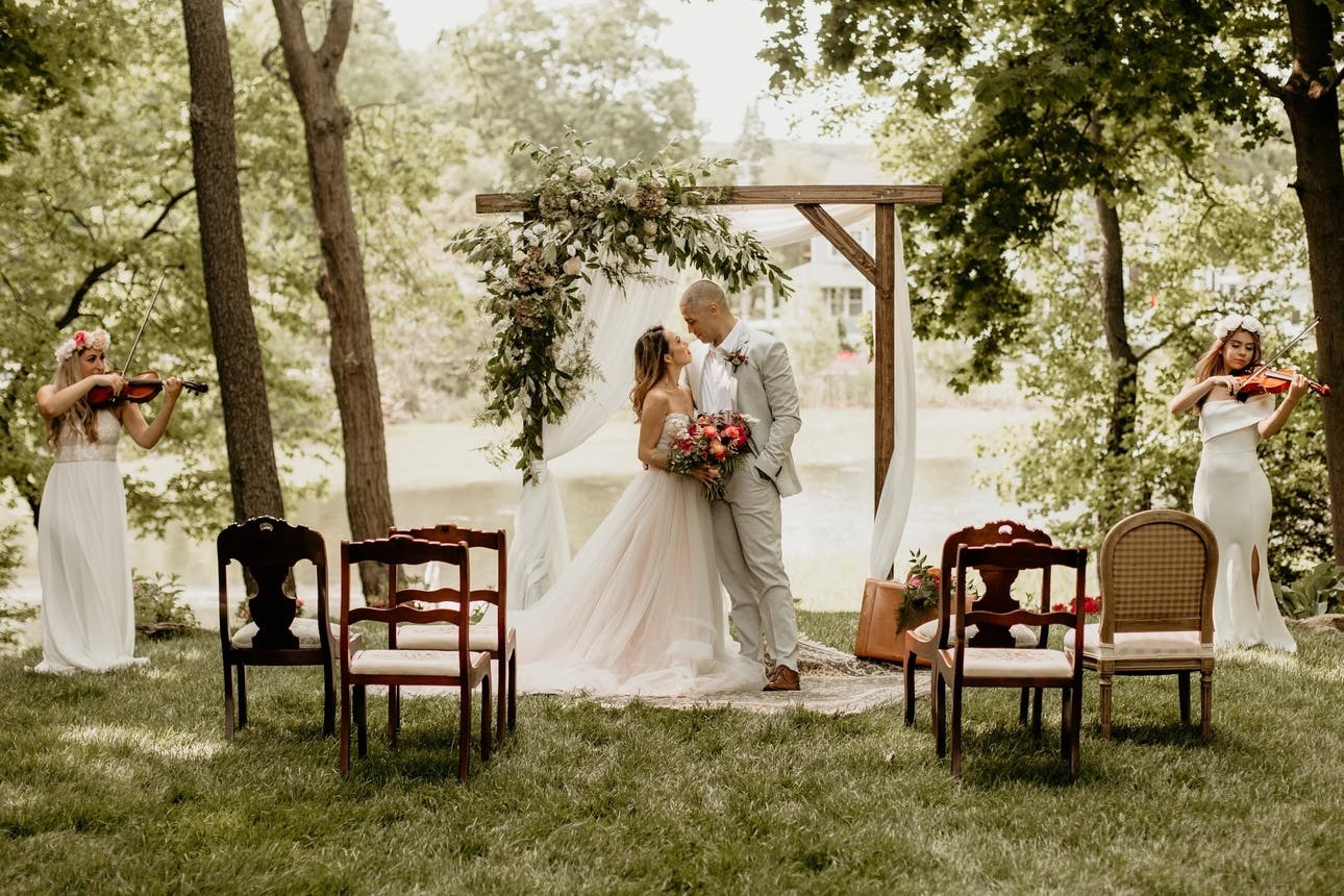 Couple stands beneath a wooden wedding arch at their intimate outdoor wedding ceremony with six chairs