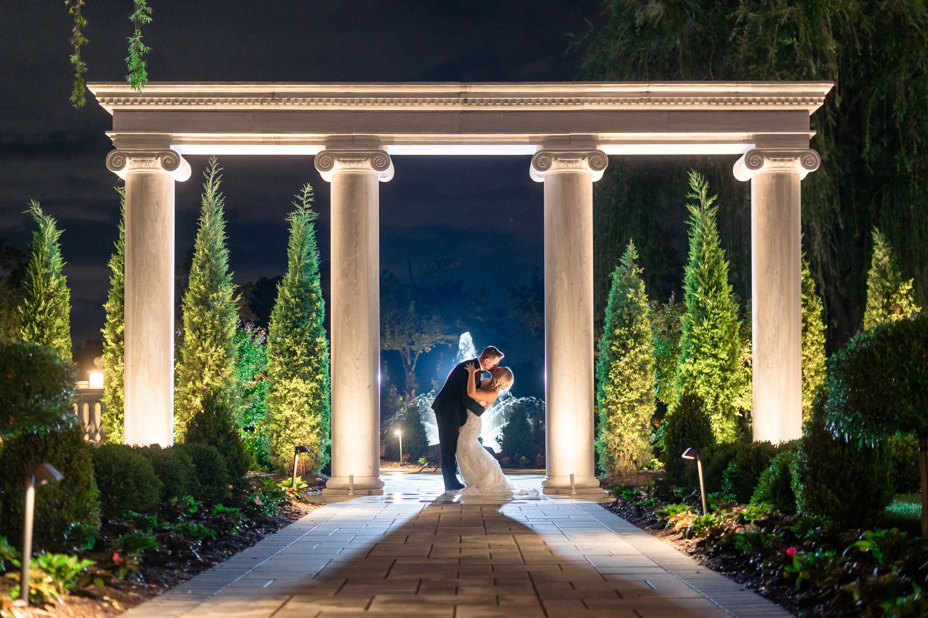 Bride and groom kissing beneath illuminated columns in a formal garden at night
