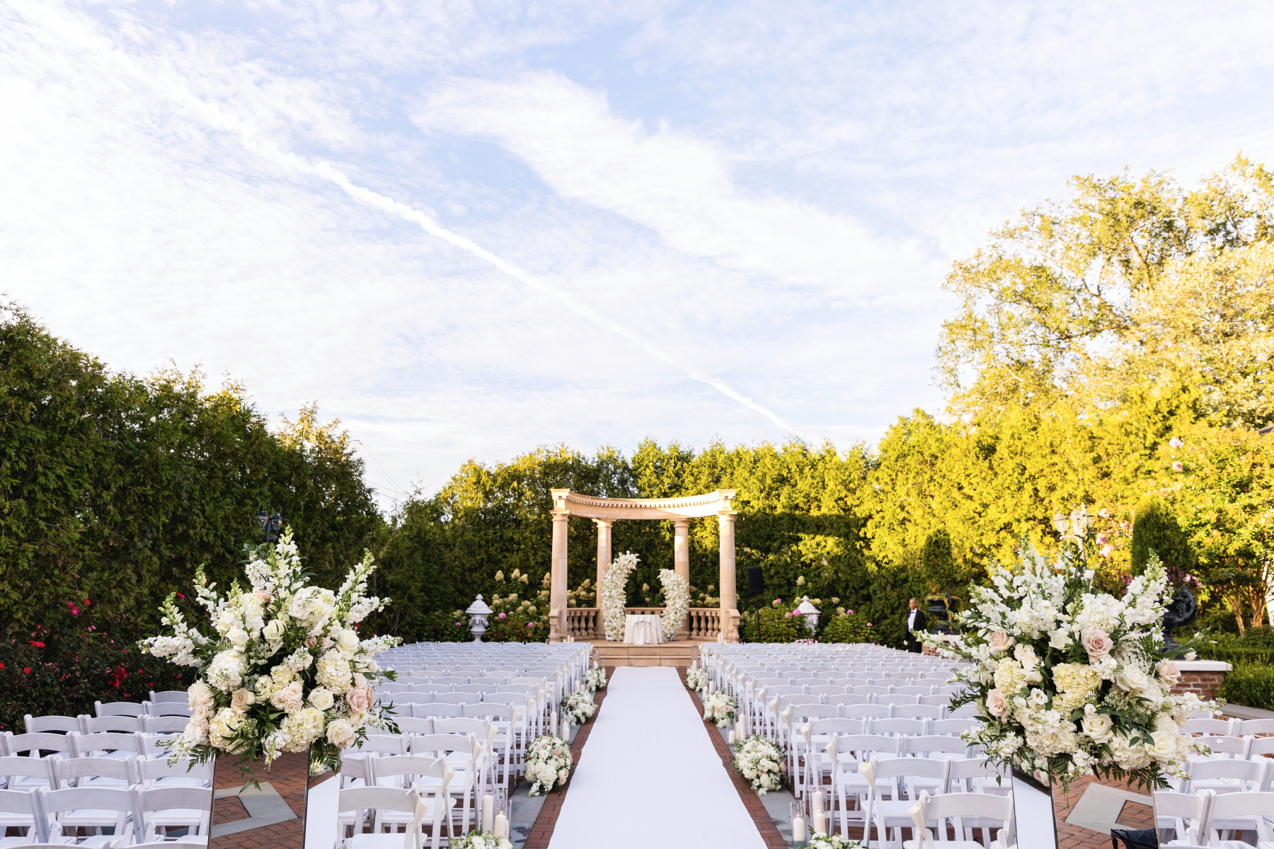 Outdoor wedding ceremony with white floral arrangements, aisle seating, and a garden pergola at The Rockleigh