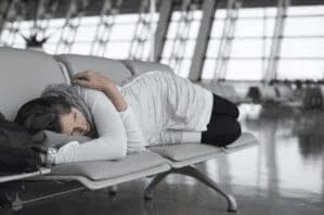A person lies curled up and sleeping on a row of empty airport seats, using a bag as a pillow. The airport terminal appears spacious and quiet in the background.