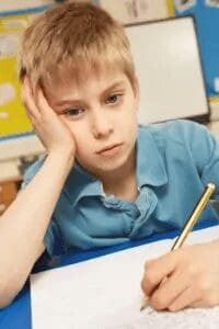 A young boy with blond hair, wearing a blue shirt, rests his head on one hand and looks bored while writing with a pencil at a desk in a classroom.