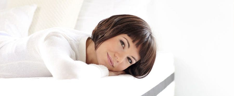 A woman with short brown hair and bangs is lying on her side on a white bed, smiling softly and resting her head on her hand, radiating a sense of calm that reflects good sleep health. The background is bright and minimalistic.