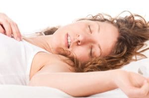 A young woman with curly hair is lying on her back in bed, eyes closed and mouth slightly open, appearing to be peacefully asleep. She is wearing a white tank top, and the background is white.