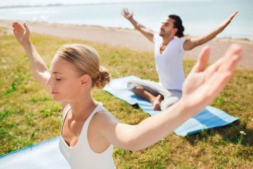 Two people practicing yoga outdoors on mats near the water, sitting cross-legged with eyes closed and arms outstretched, enjoying a peaceful, sunny day and the benefits of mindful exercise.
