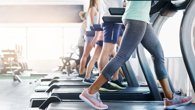 People exercising on treadmills at a gym. The focus is on the lower bodies and legs of four individuals in athletic clothing and sneakers, working out side by side in a bright, modern setting.
