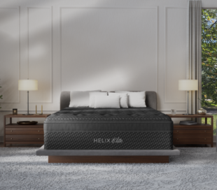 A modern bedroom features a gray Helix Elite mattress on a platform bed, flanked by wooden nightstands with matching lamps. A large window lets in natural light, and a green plant sits on the left side of the room.