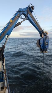 Crane that deposits and retrieves Jason from the water with cable in the water at sunset. M. Talbot, University of Washington, V26.