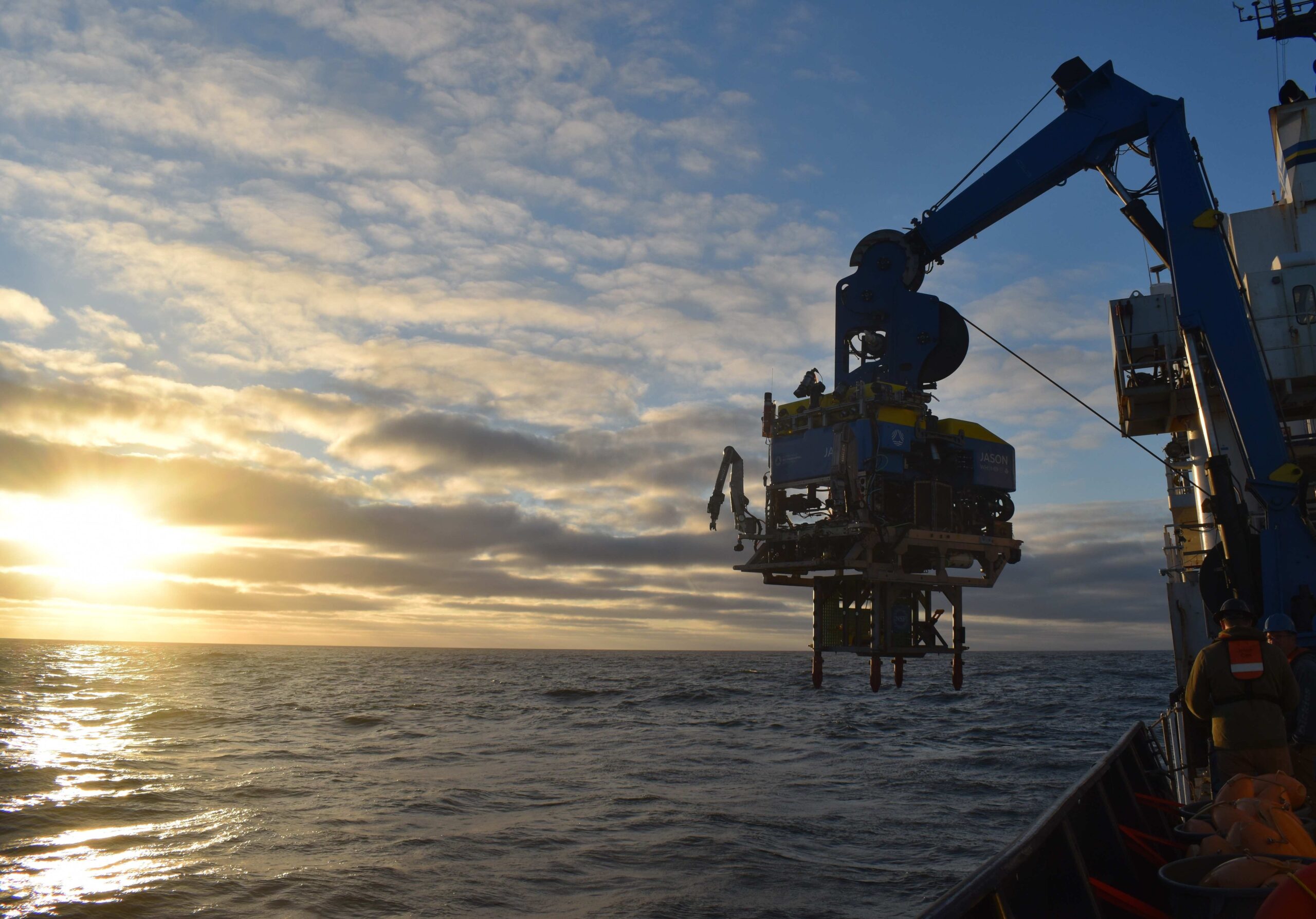 A remotely operated vehicle with a metal frame latched below is moved over the side of a ship while the sunsets into the ocean in the background.