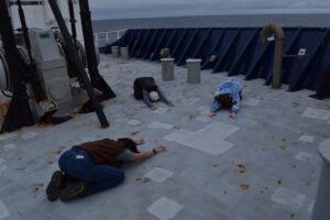 Three people do child's pose on the gray deck of a ship's bow. Ship's equipment is in the background and you can see the ocean over the ship's rail.