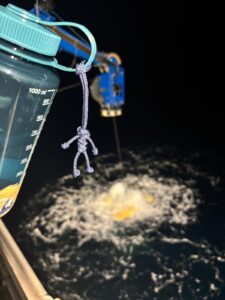 A blue rope man hanging from a turquoise water bottle. In the background a blue crane winches a yellow robot out of the water.