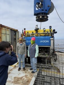 Dr. Kelley wearing a fish mask and pink hard hat, and Dr. Bigham wearing a lizard head mask, pose in front of the ROV Jason. Credit: A. Chang, University of Washington; V26.