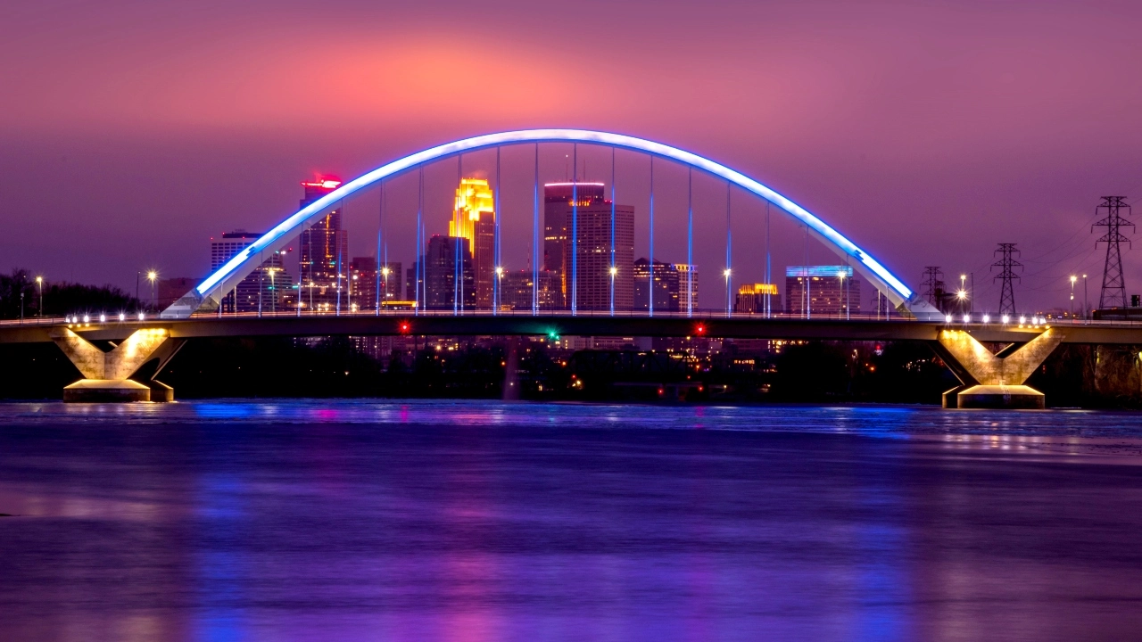 Image of the Lowry Avenue Bridge, lighted blue at dusk, with Minneapolis skyline in the background