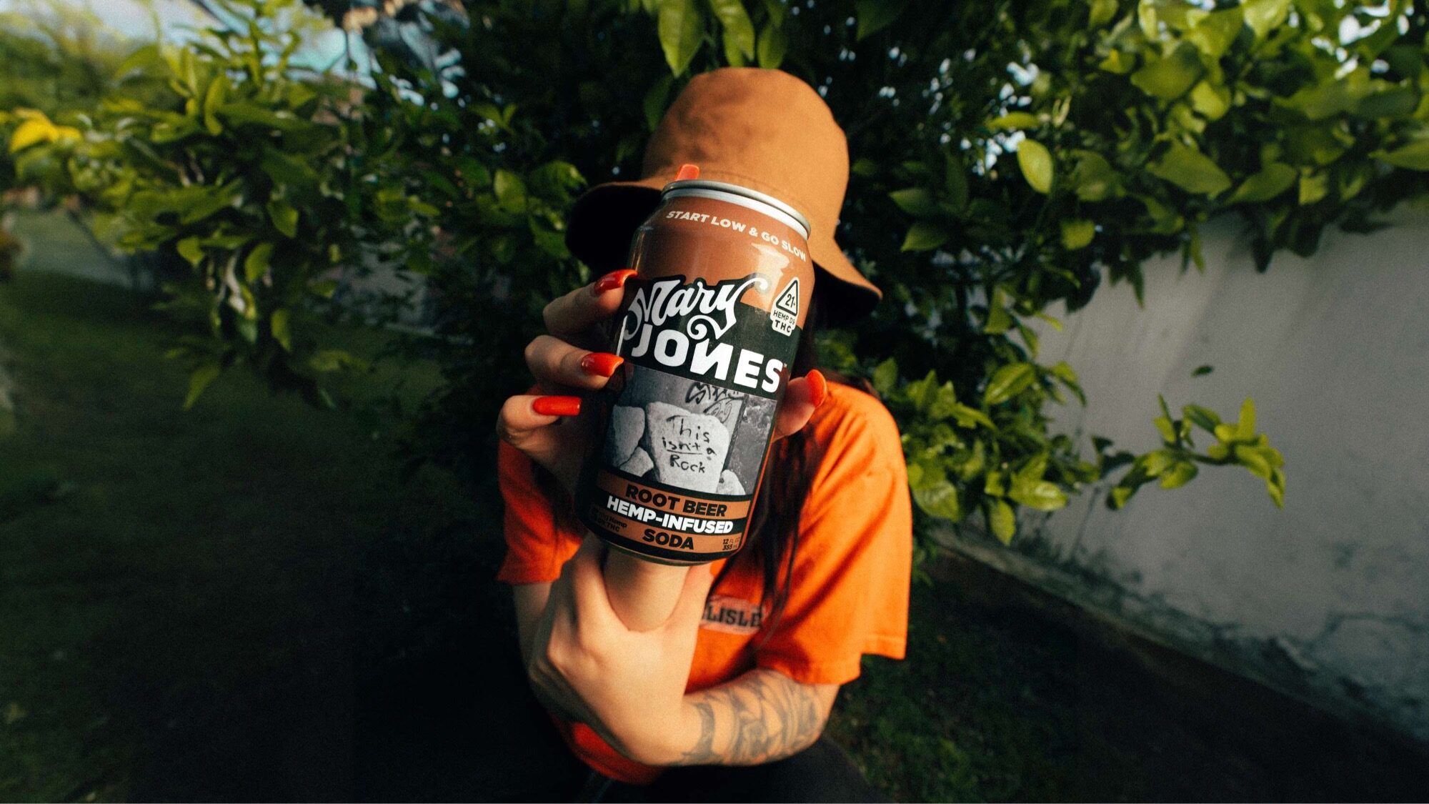 Person in an orange shirt and hat holds a can of Mary Jones root beer hemp-infused soda in front of their face, with greenery and a white wall in the background.
