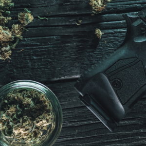 A small glass jar of legal cannabis flower sits on a table next to a black handgun