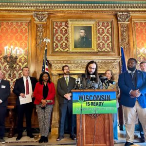 A group of people stand behind a podium with a Wisconsin Is Ready To Grow Our Future sign, speaking at a press event in an ornate room with a portrait on the wall.