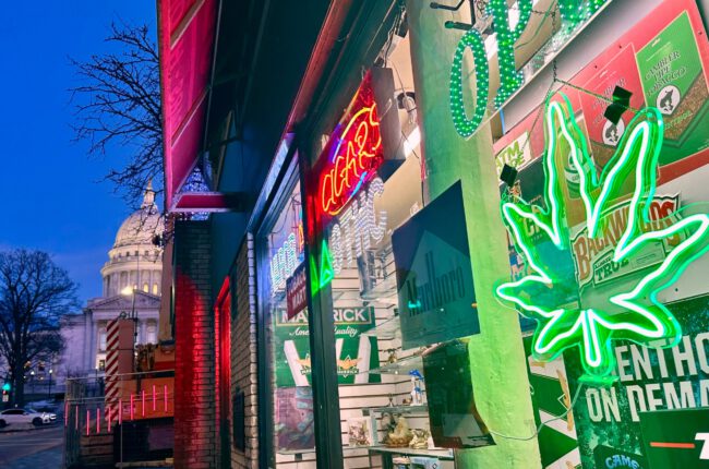 Storefront with neon signs, including an open sign and a green cannabis leaf, at dusk; a capitol building is visible in the background across the street.