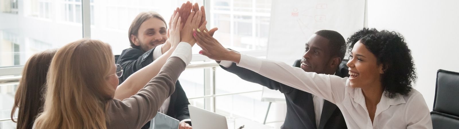 Happy business meeting with five smiling, diverse professionals high-fiving signifying collaboration.
