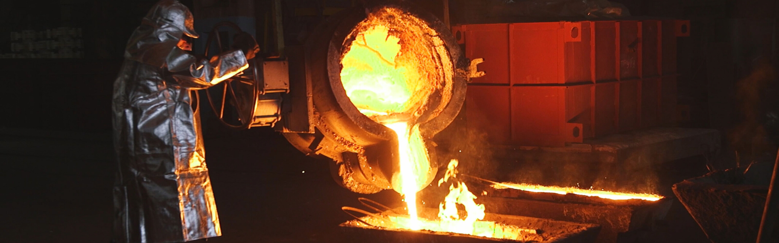 Smelter employee in a silver protective hazmat suit pouring molten gold from a heavy industrial pot into a container on the ground.
